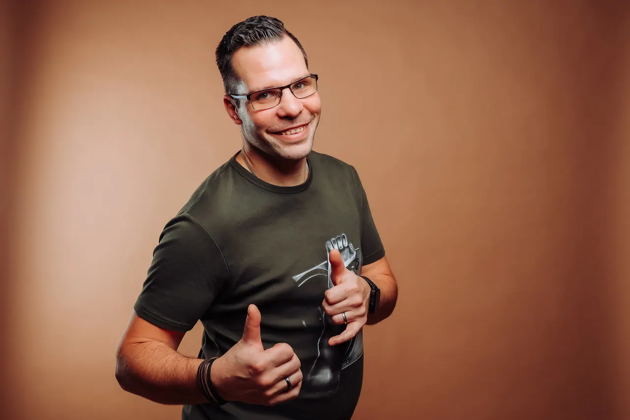 Dominik Hans lacht und zeigt beide Daumen nach oben, brauner Studiohintergrund
