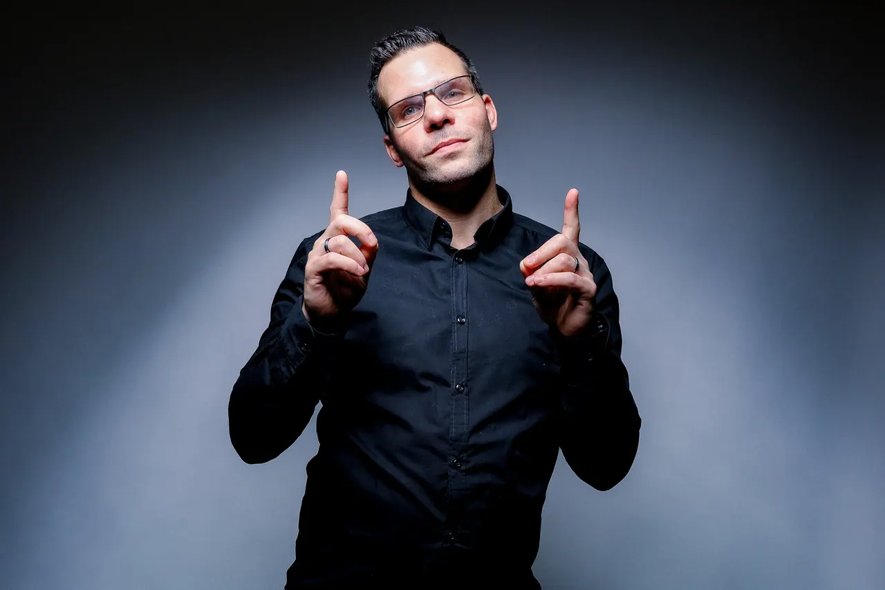 Dominik Hans im schwarzen Hemd vor blaugrauem Licht, beide Zeigefinger nach oben